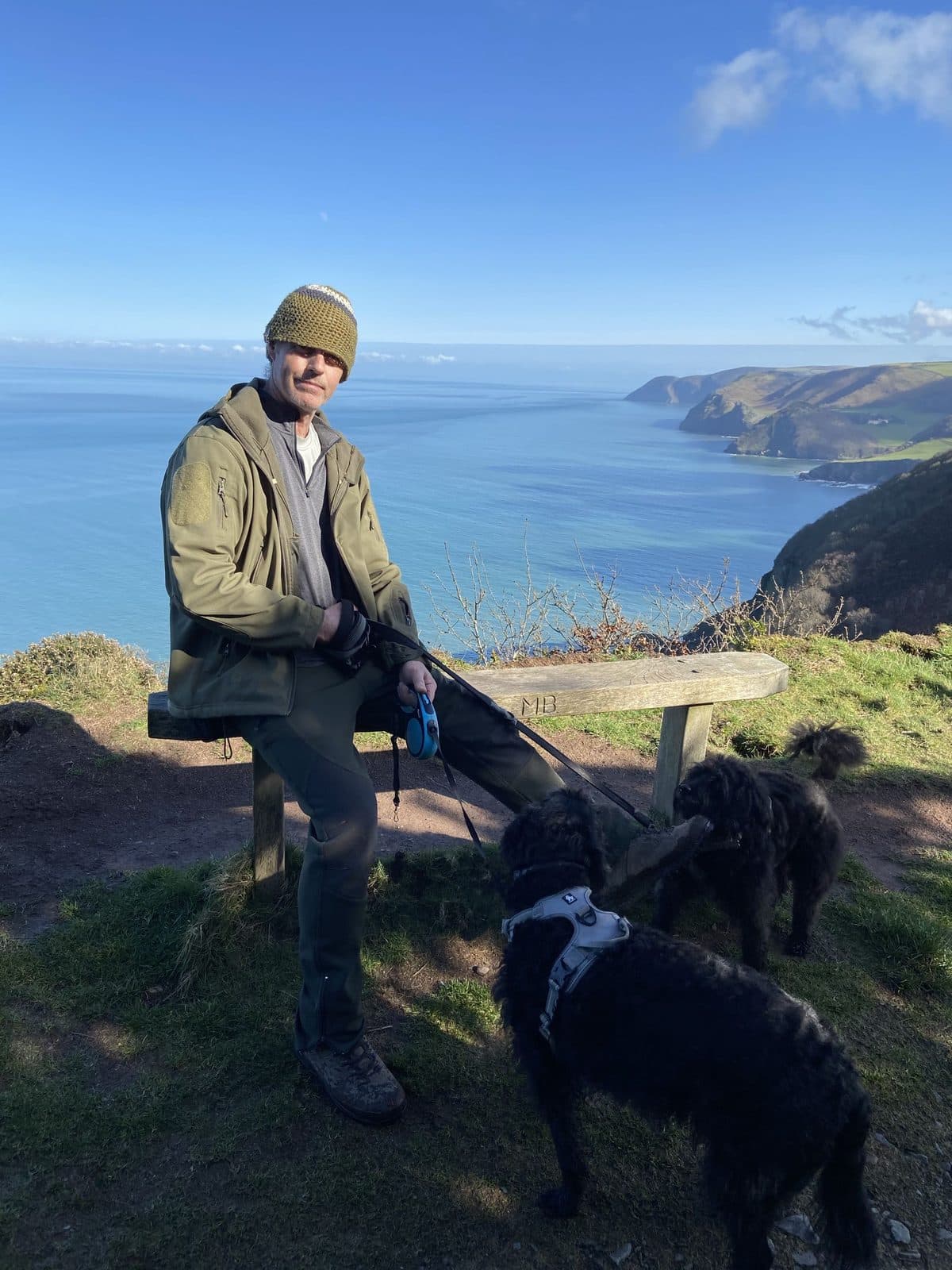 Pete Carling sitting on a bench on the South West Coast Path above the sea, two black dogs beside him