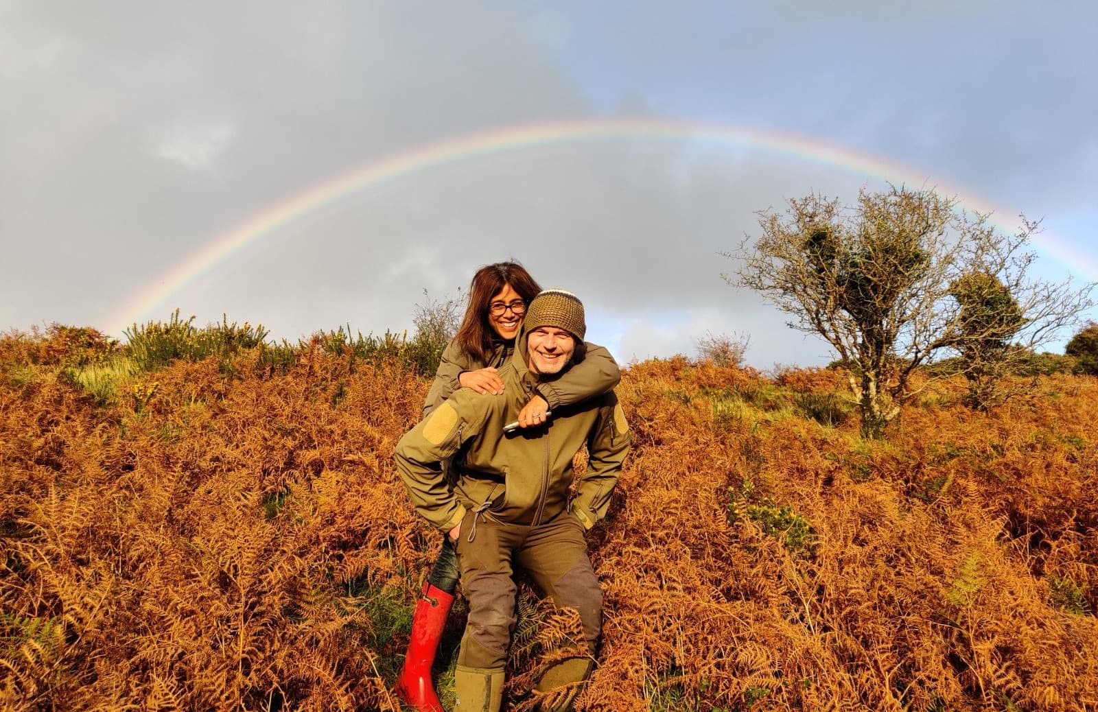 Pete and Nicole laughing together on the moor above the Bristol Channel, a full rainbow arcing over the hillside