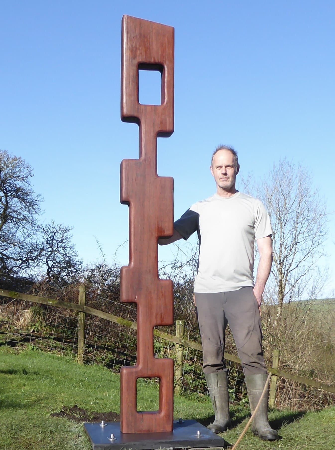Pete Carling standing beside one of his hand-made timber sculptures in a Devon field, under a clear blue sky