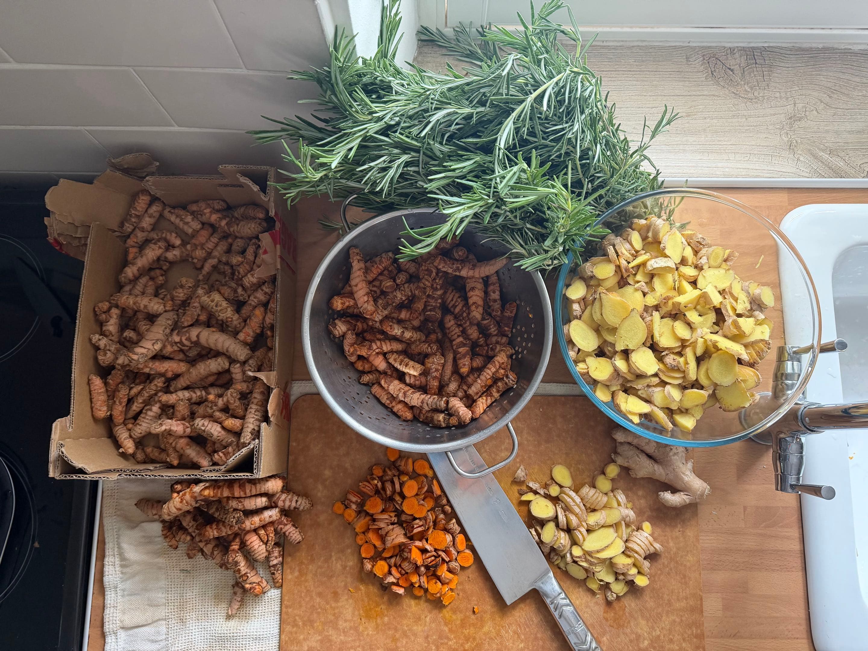 Fresh turmeric and ginger being prepared by hand on a Devon kitchen counter, with rosemary and a colander of turmeric root